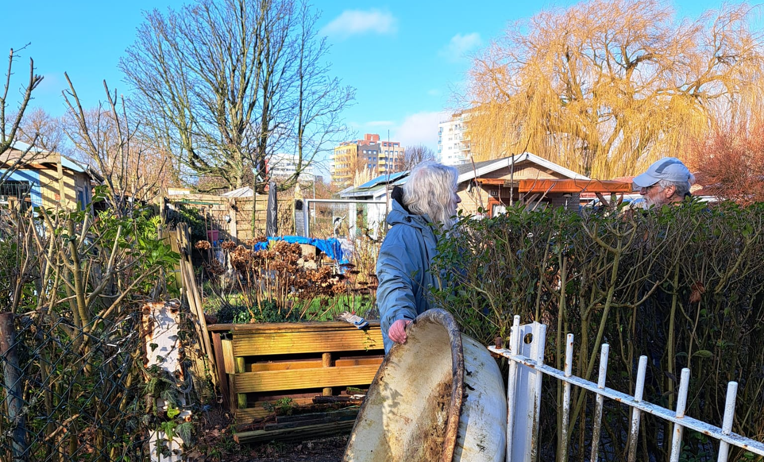 Een man draagt een metalen voorwerp door een poort in een tuin; een andere persoon is zichtbaar bij een heg op een zonnige dag.