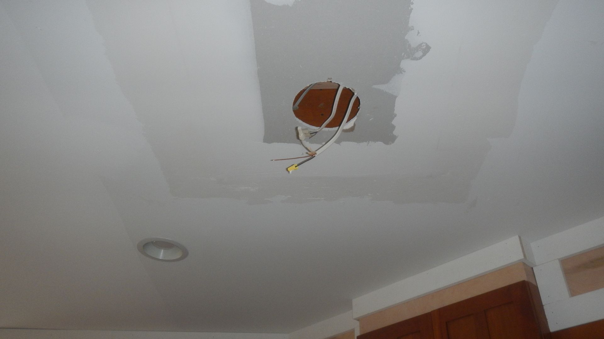 Ceiling with a hole for wiring, surrounded by patched drywall. Brown cabinets are visible below.