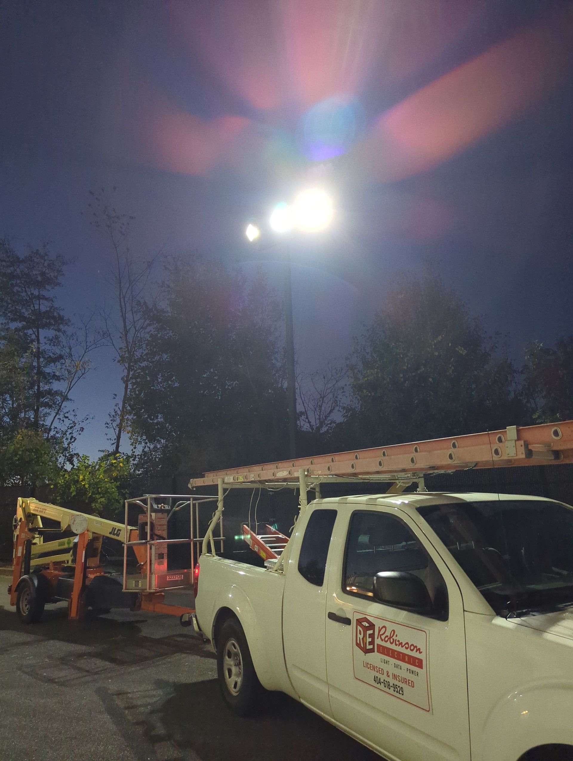A white work truck and an orange lift platform next to a tall light pole at night.