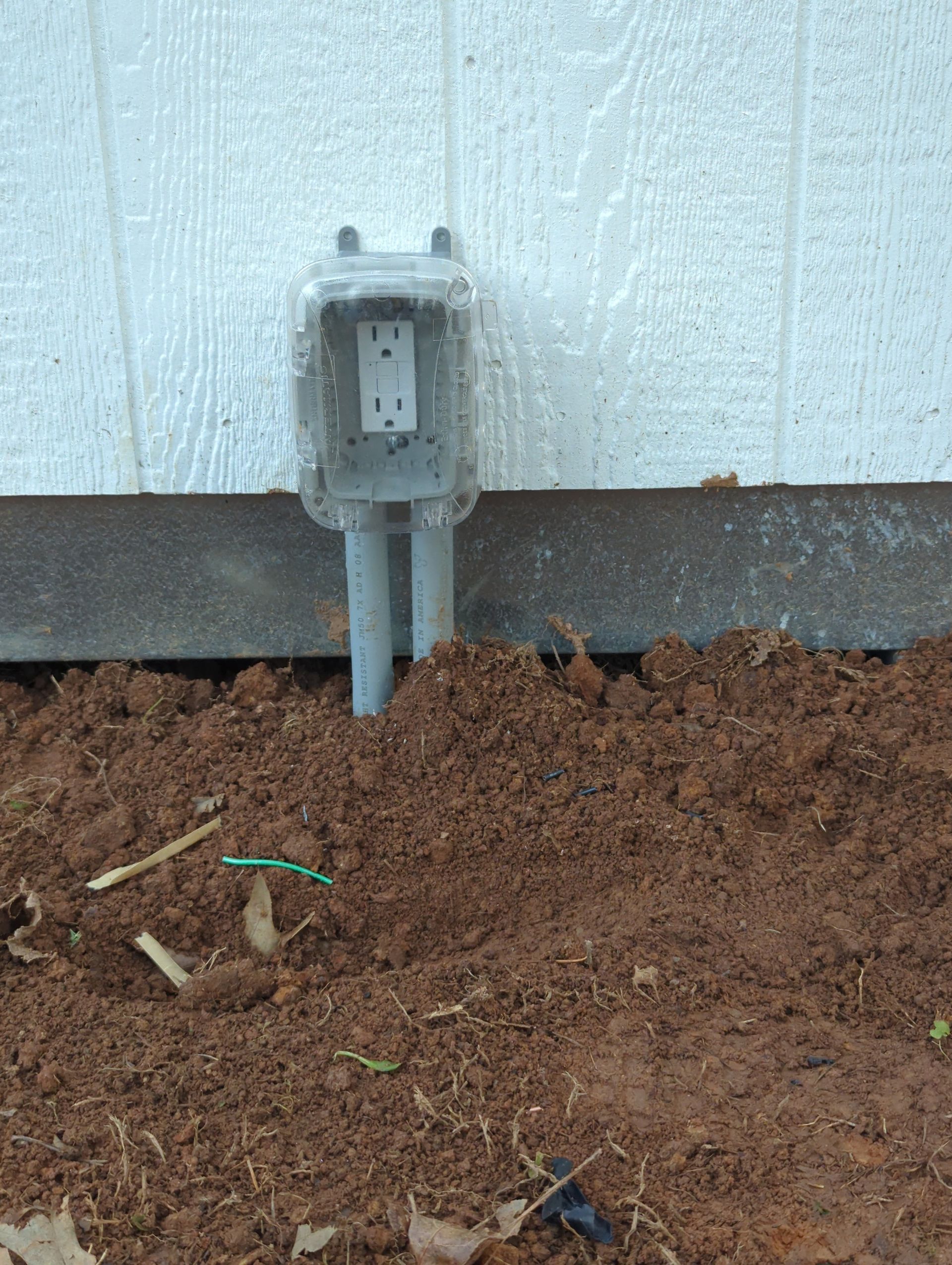 Outdoor electrical outlet with a protective cover installed on a white building exterior, in a bed of dirt.