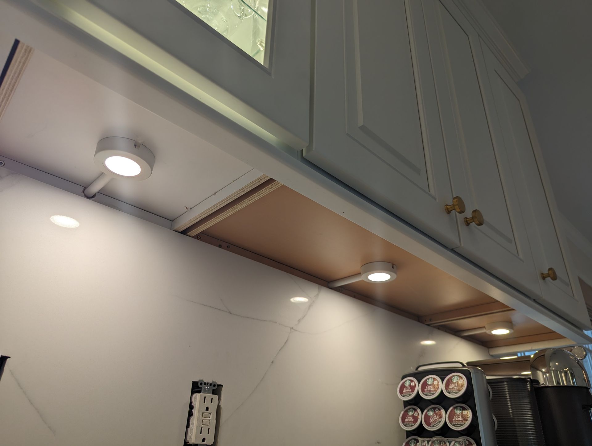 Low-profile LED puck lights installed under white kitchen cabinets, illuminating a white marble backsplash and wall outlet.