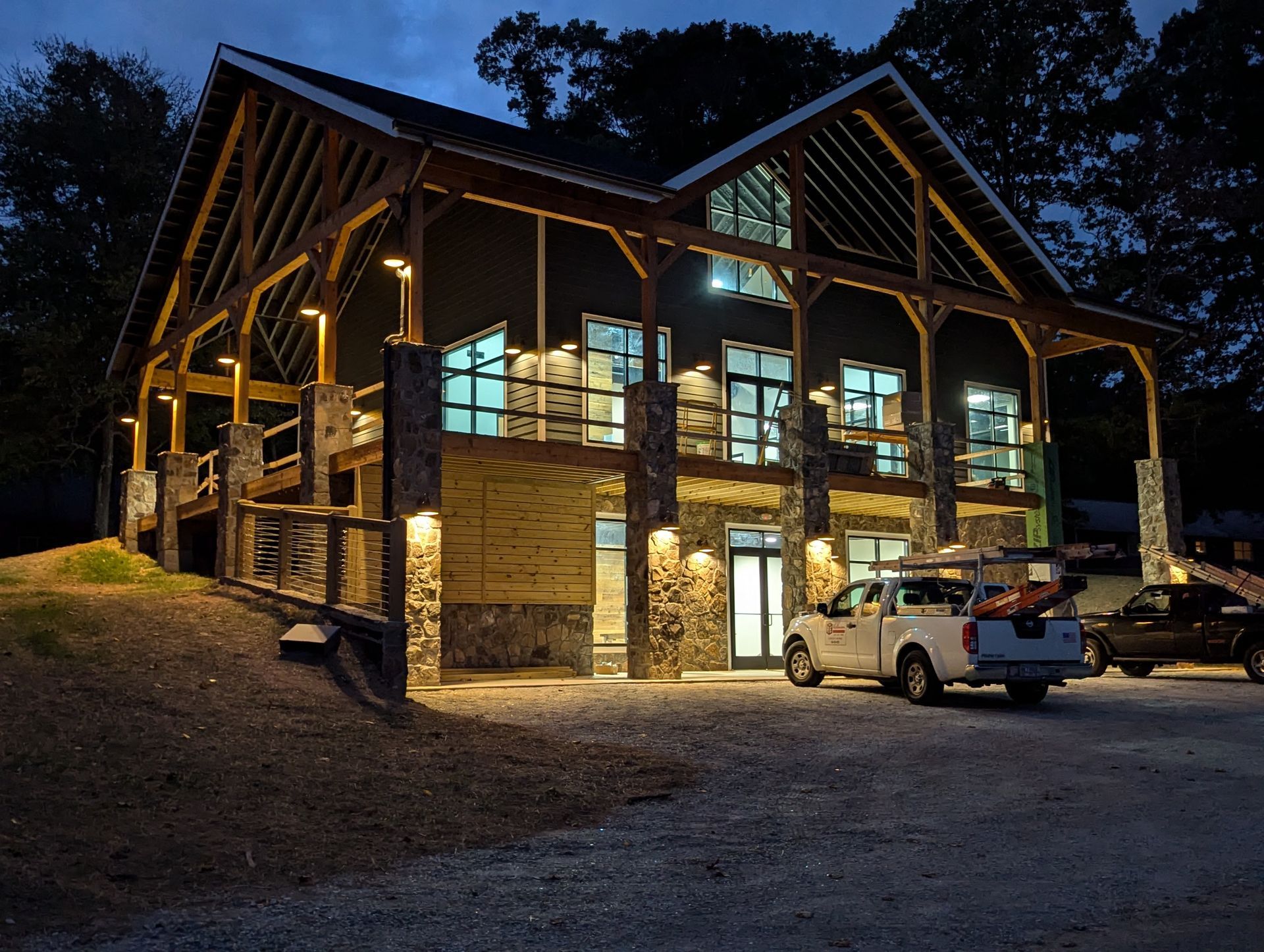 Two-story timber-frame building with stone accents, illuminated at twilight, featuring a deck and parked pickup trucks.