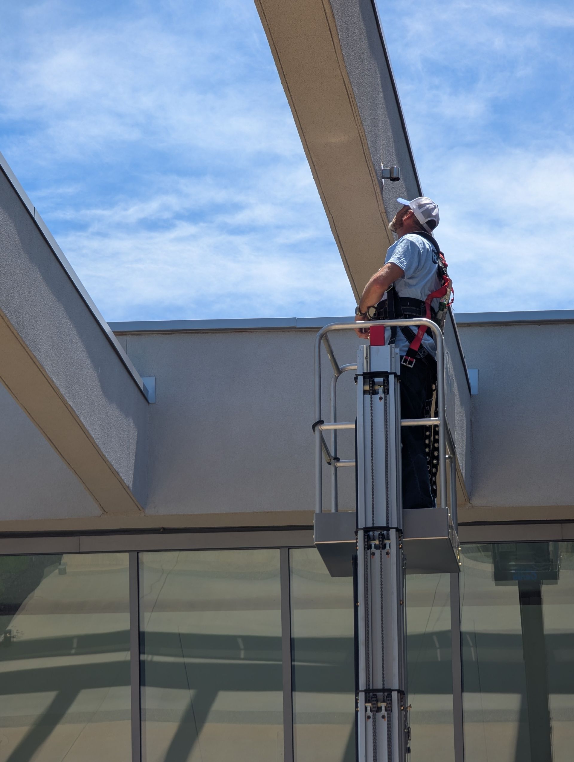 Worker on a lift inspecting an overhead beam, wearing a hard hat and safety harness. Blue sky in the background.