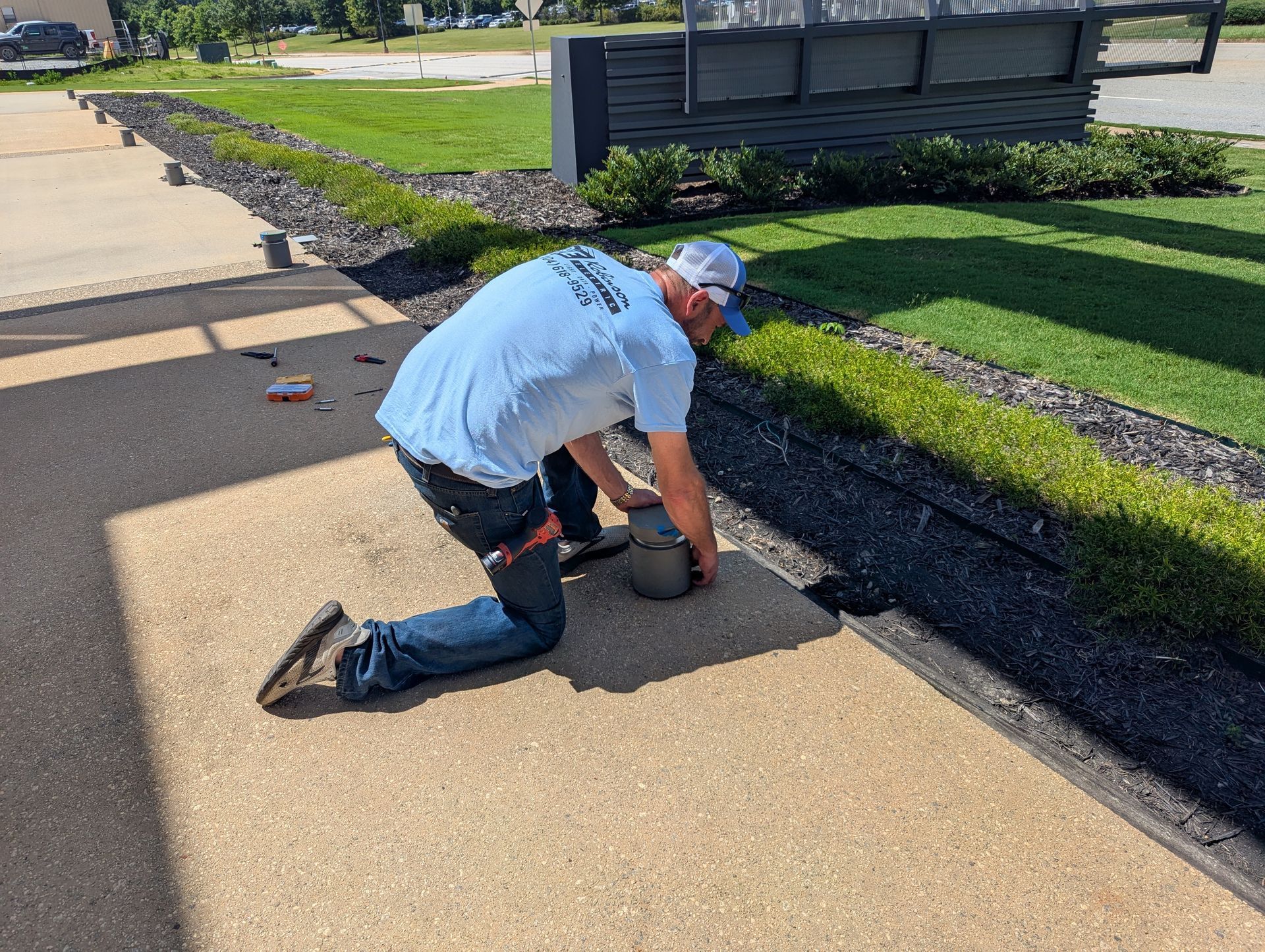 Man kneeling, working on pavement near landscaping. He's wearing a light blue shirt and jeans. Outdoors.