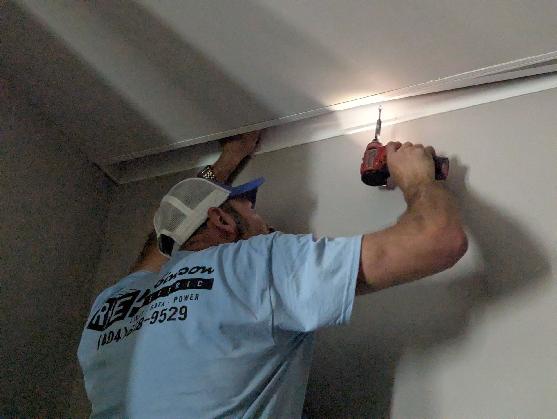 Person in blue shirt using a power drill to install molding on a white ceiling.