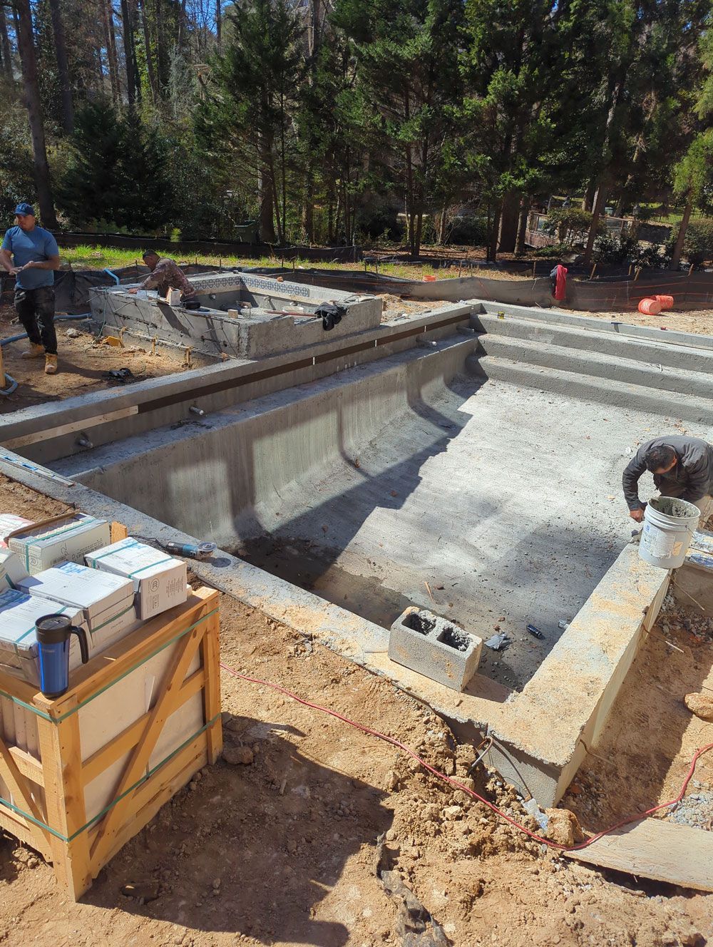 Pool construction site; concrete pool shell with steps; workers, wooden crate of tiles, and surrounding dirt.