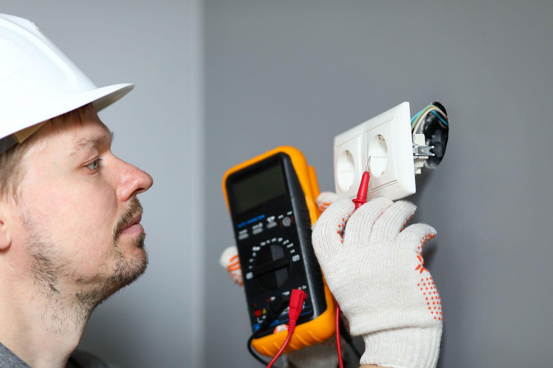 An electrician in a hard hat and protective gloves uses a digital multimeter to test a wall-mounted electrical outlet.