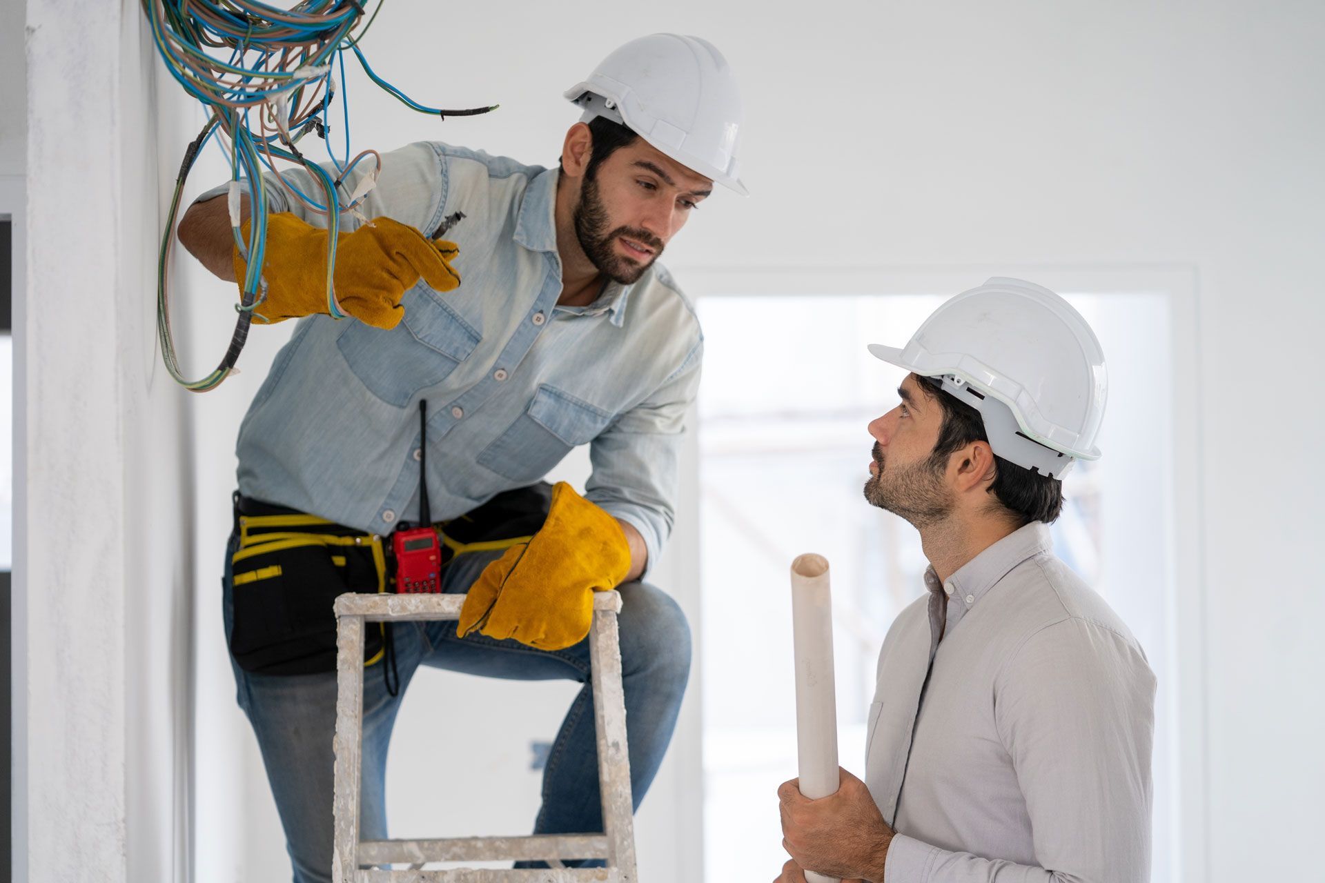 Two professionals in hard hats discuss electrical wiring at a construction site; one stands on a ladder, pointing at cables.