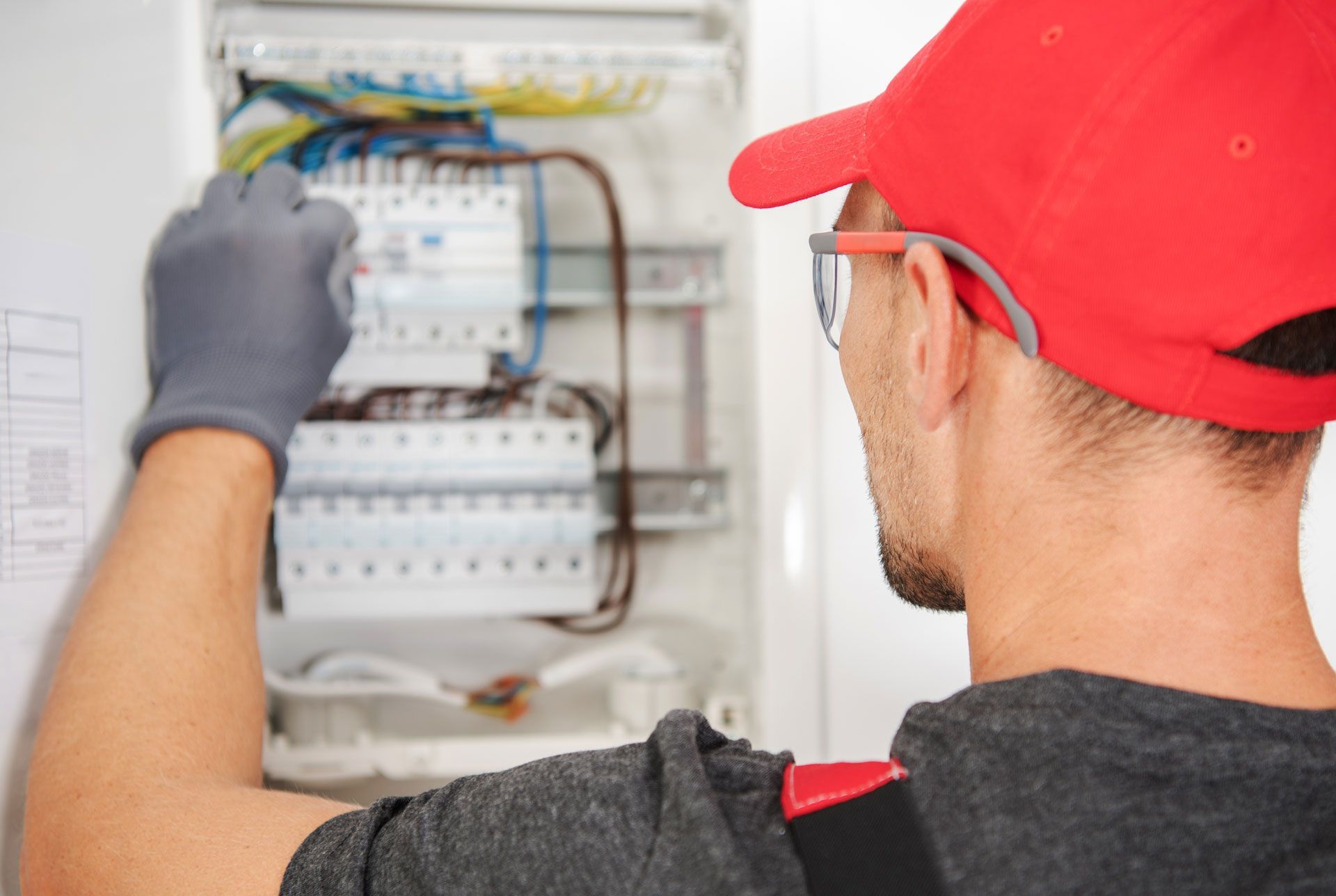 Electrician in a red cap and safety glasses working on an open electrical panel with wires.