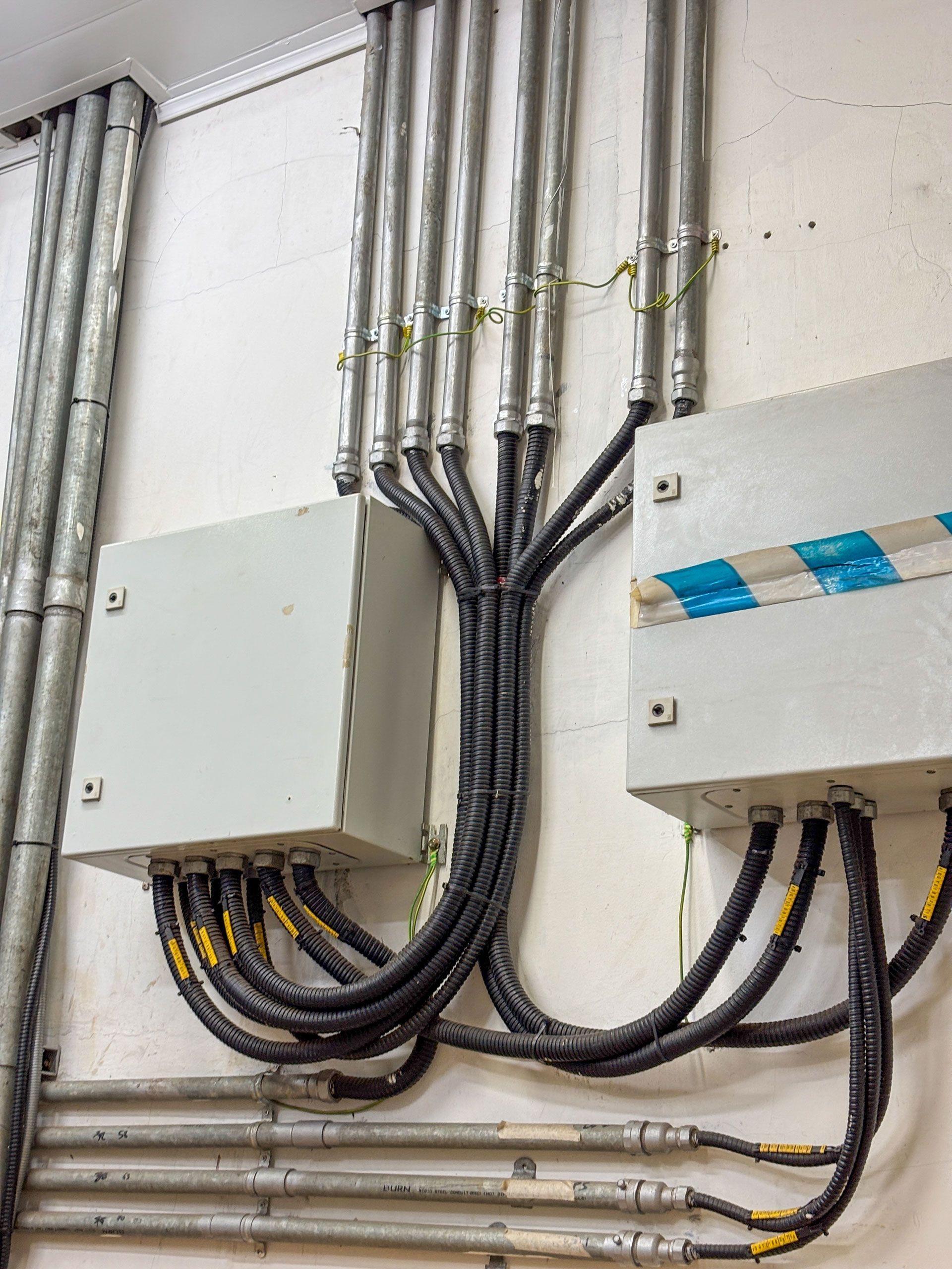 Electrical conduit pipes mounted on a white wall connect to two gray rectangular junction boxes with black power cables.
