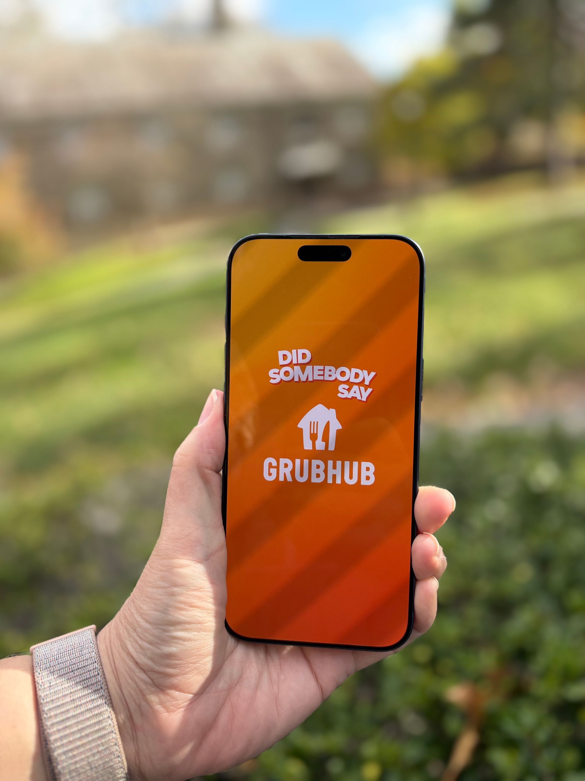 Hand holding phone displaying Grubhub logo. Outdoor setting with green grass and a building.