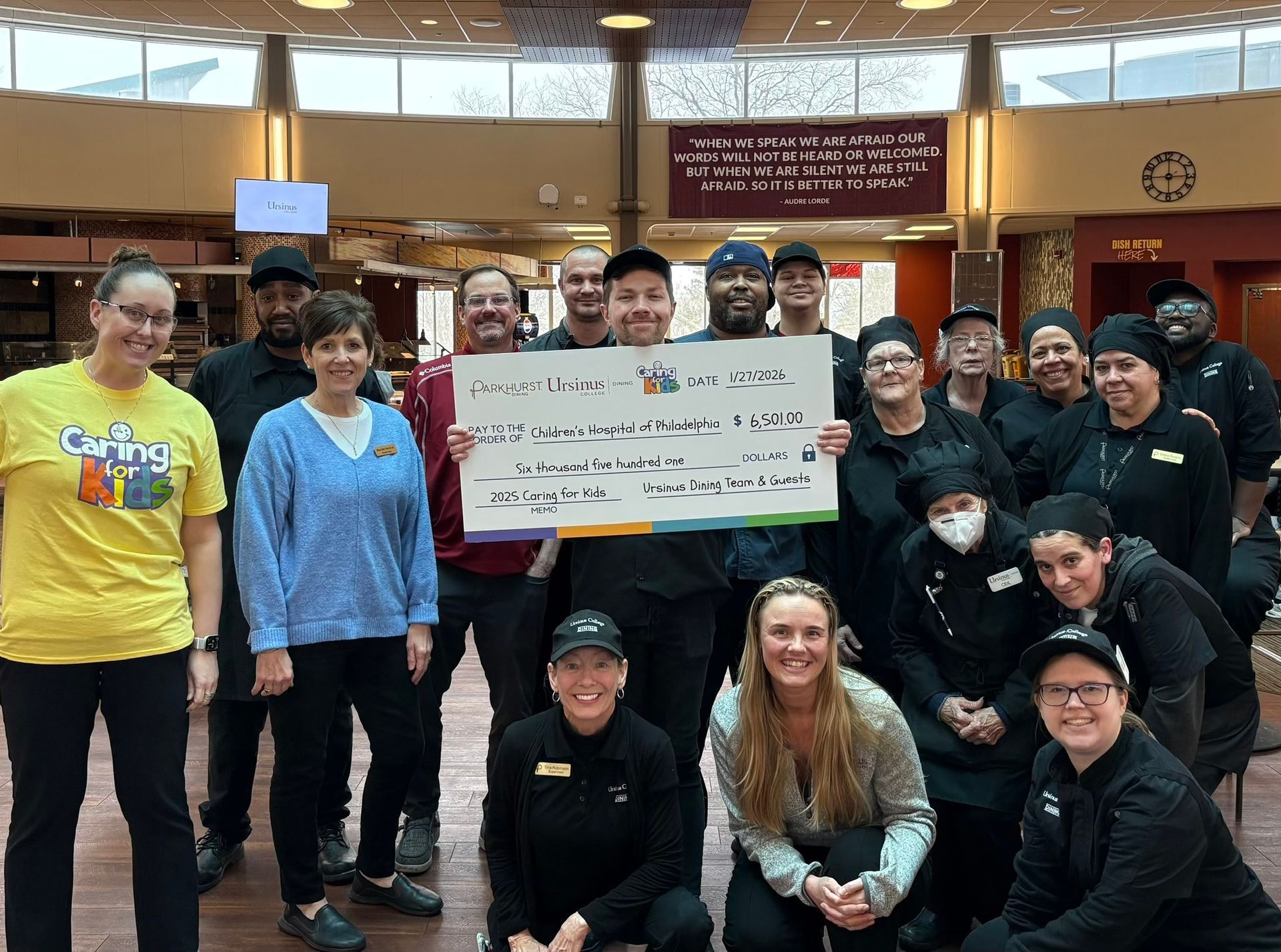 A group of food service workers in uniform smiles while holding a large sign, standing in a brightly lit dining hall.
