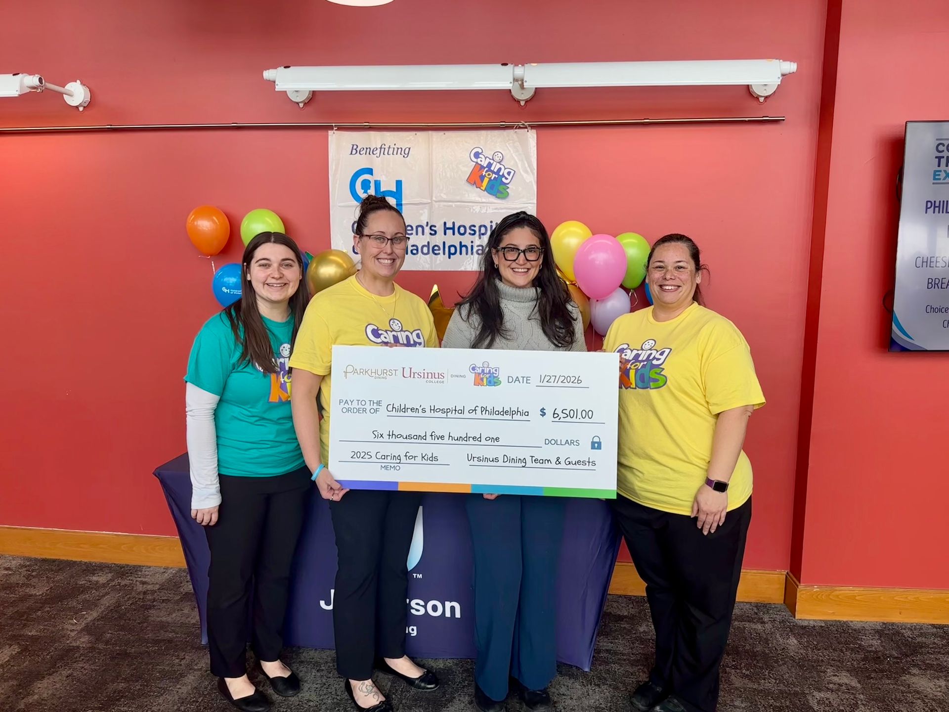 Four people smile while holding a large donation check for Children's Hospital of Philadelphia in a room with balloons.