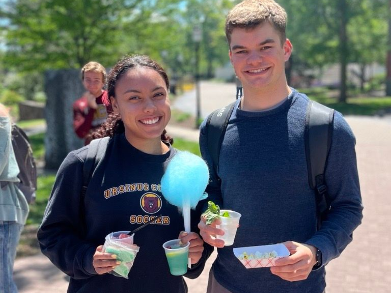 Two people smiling, holding cotton candy, snacks, and drinks outdoors on a sunny day.