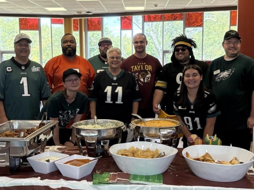 People at a football party, serving food from a decorated table. Many wear team jerseys and smiles.