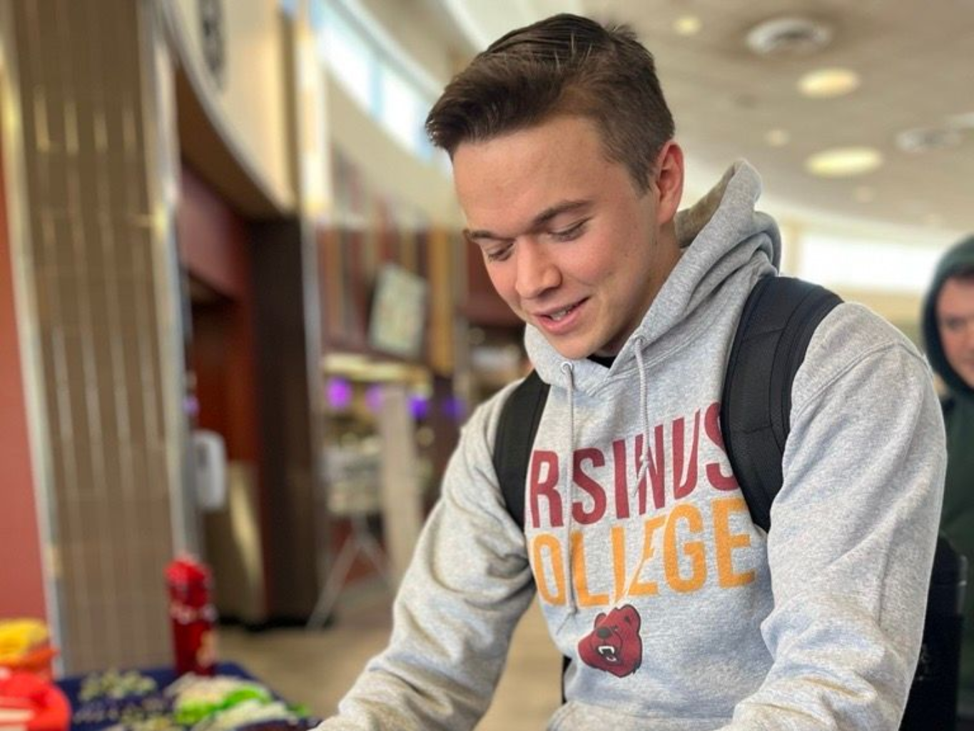 Man scoops cereal from bowls at a table. He wears a grey hoodie with a school logo and a backpack.