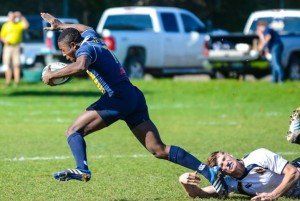 A man is running with a rugby ball while another man is laying on the ground.