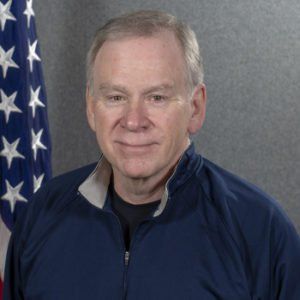 A man in a blue jacket stands in front of an american flag