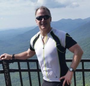 A man wearing sunglasses is leaning on a railing with mountains in the background