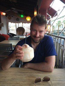 A man is sitting at a table drinking from a coconut.