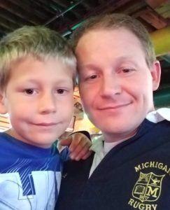 A man and a boy are posing for a picture and the man is wearing a michigan rugby jacket