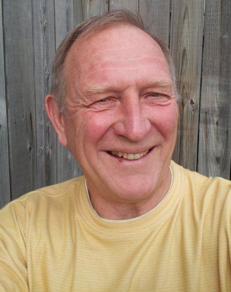 A man in a yellow shirt is smiling in front of a wooden fence