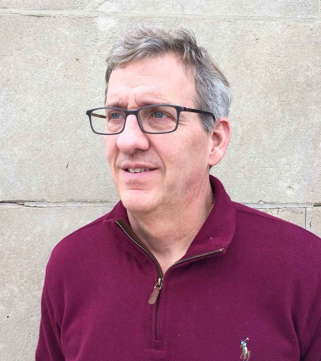A man wearing glasses and a maroon sweater is standing in front of a brick wall.