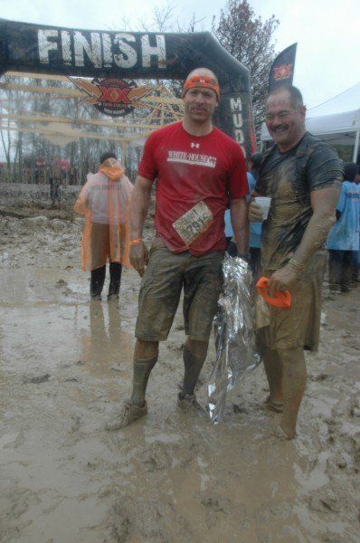 Two muddy men standing in front of a finish sign