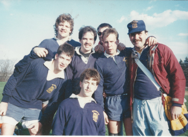 A group of men are posing for a picture with one wearing a shirt that says ' rugby ' on it