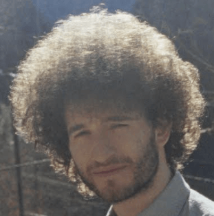 A man with curly hair and a beard looks at the camera