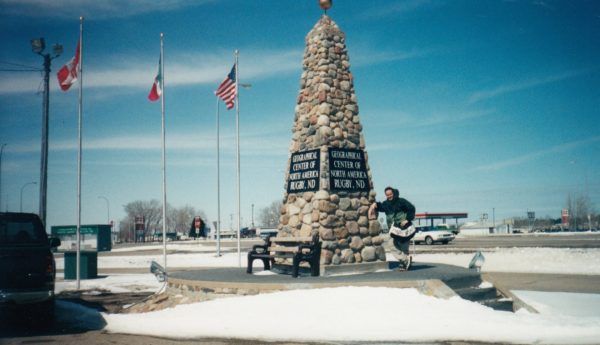 A man stands in front of a stone obelisk with flags in the background.