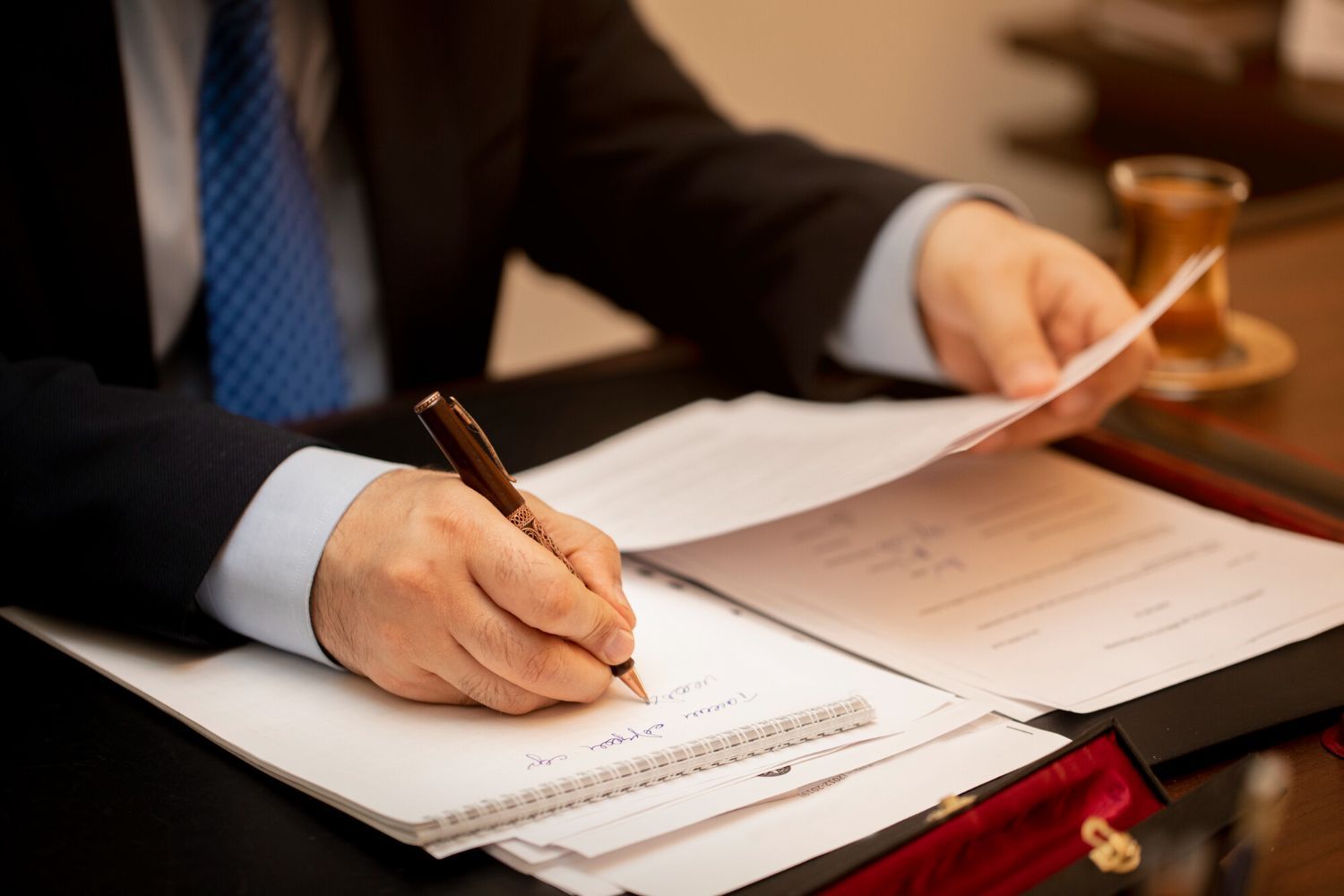 Hand in a suit signing a document at a desk with papers and a stamp