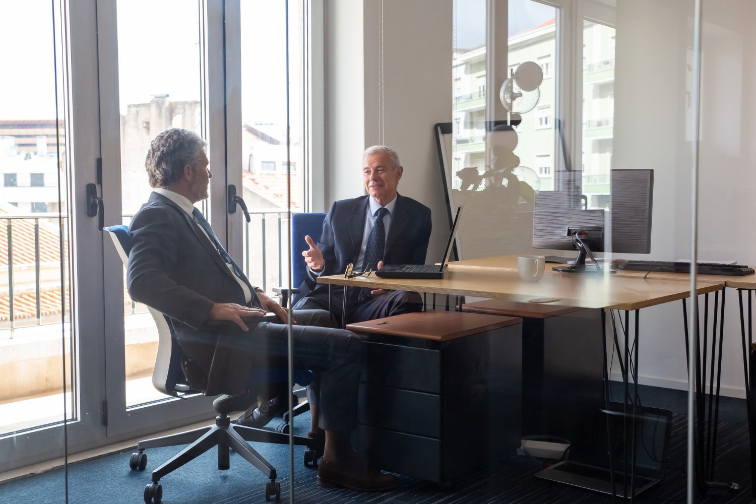 Two people talking in a glass-walled office, seated across a table with laptops and papers.