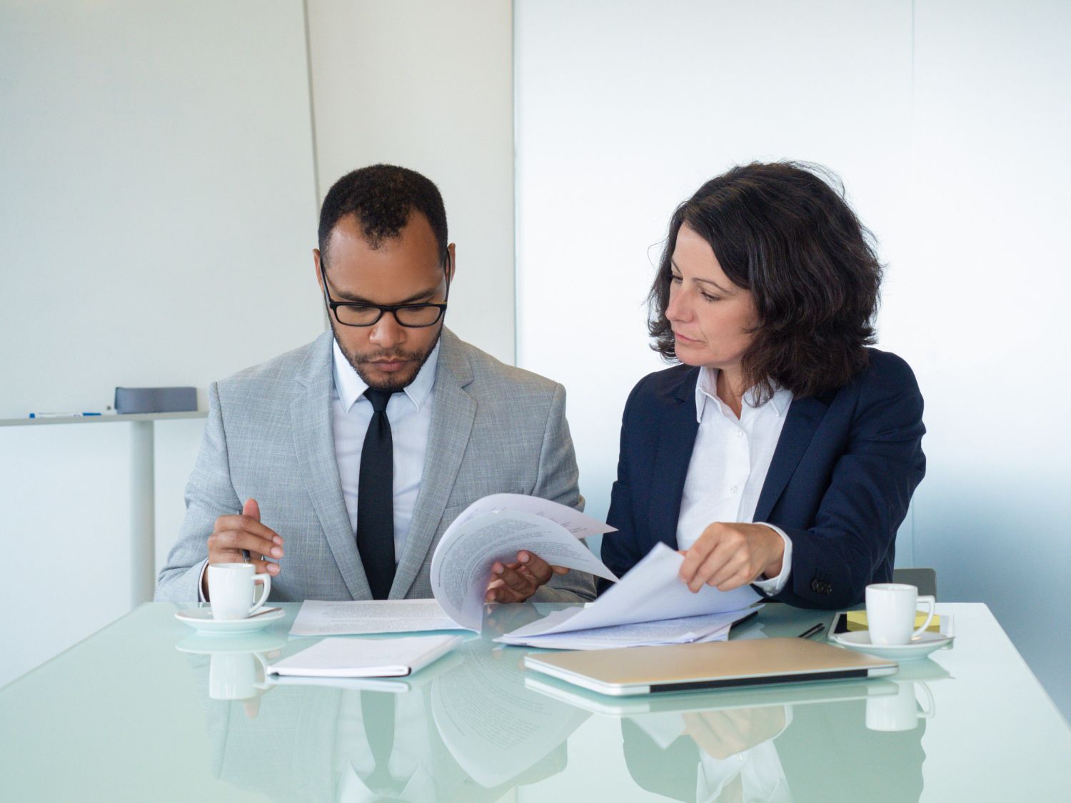 Two coworkers review documents at a glass table in a bright office.