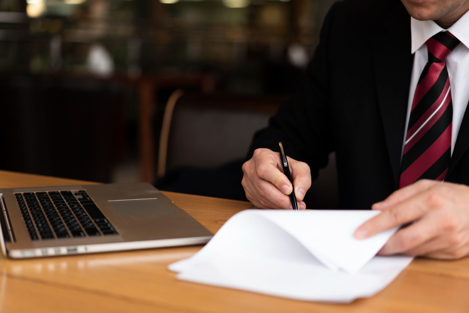 Person in a dark suit signs a document beside a laptop on a wooden desk