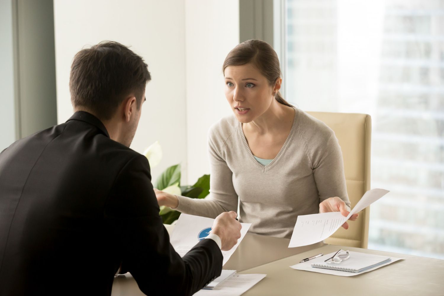 Two people talking at a desk, one holding papers in a bright office.