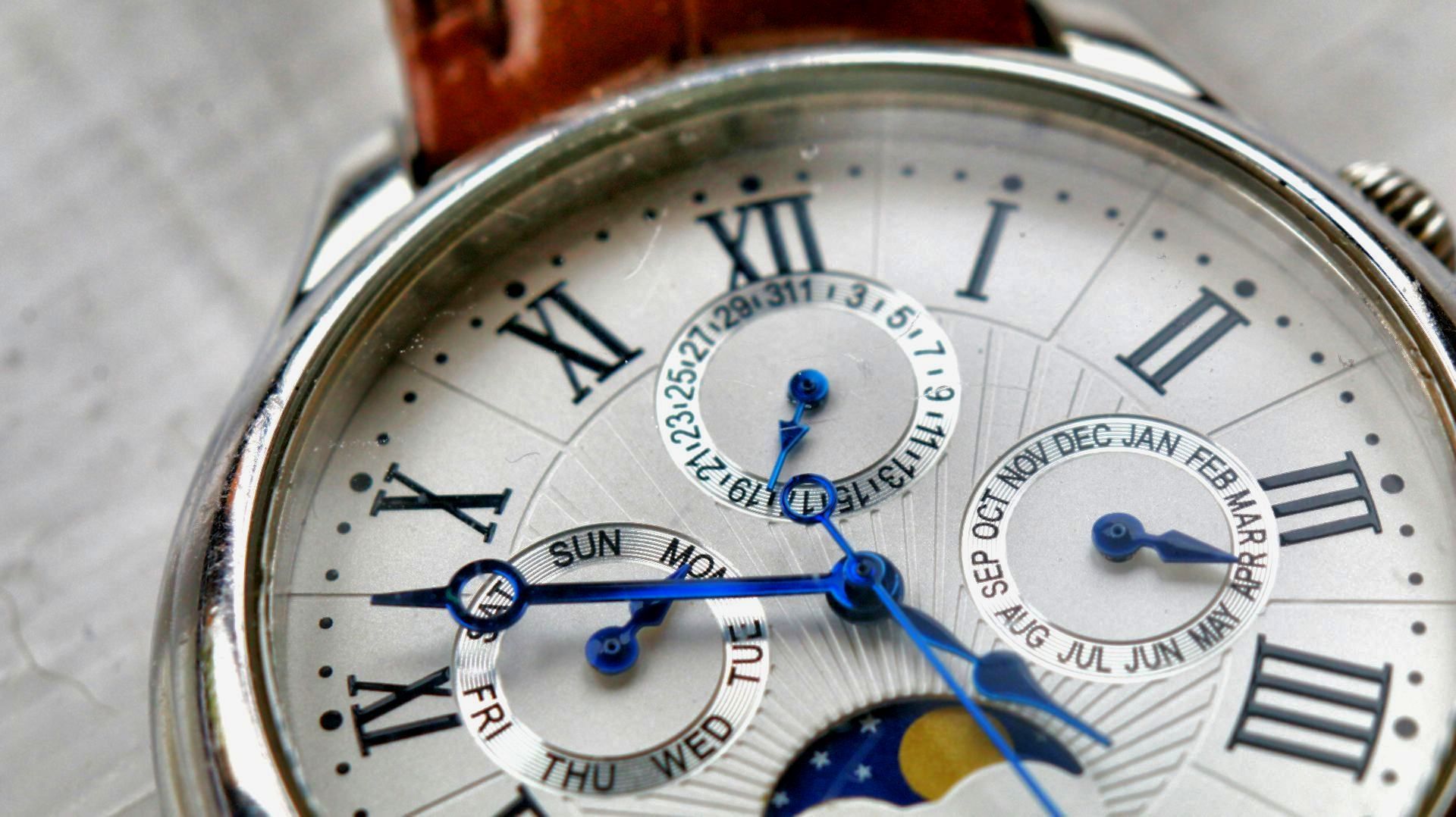 Close-up of a watch with Roman numerals, three sub-dials, and a leather band. Blue details.