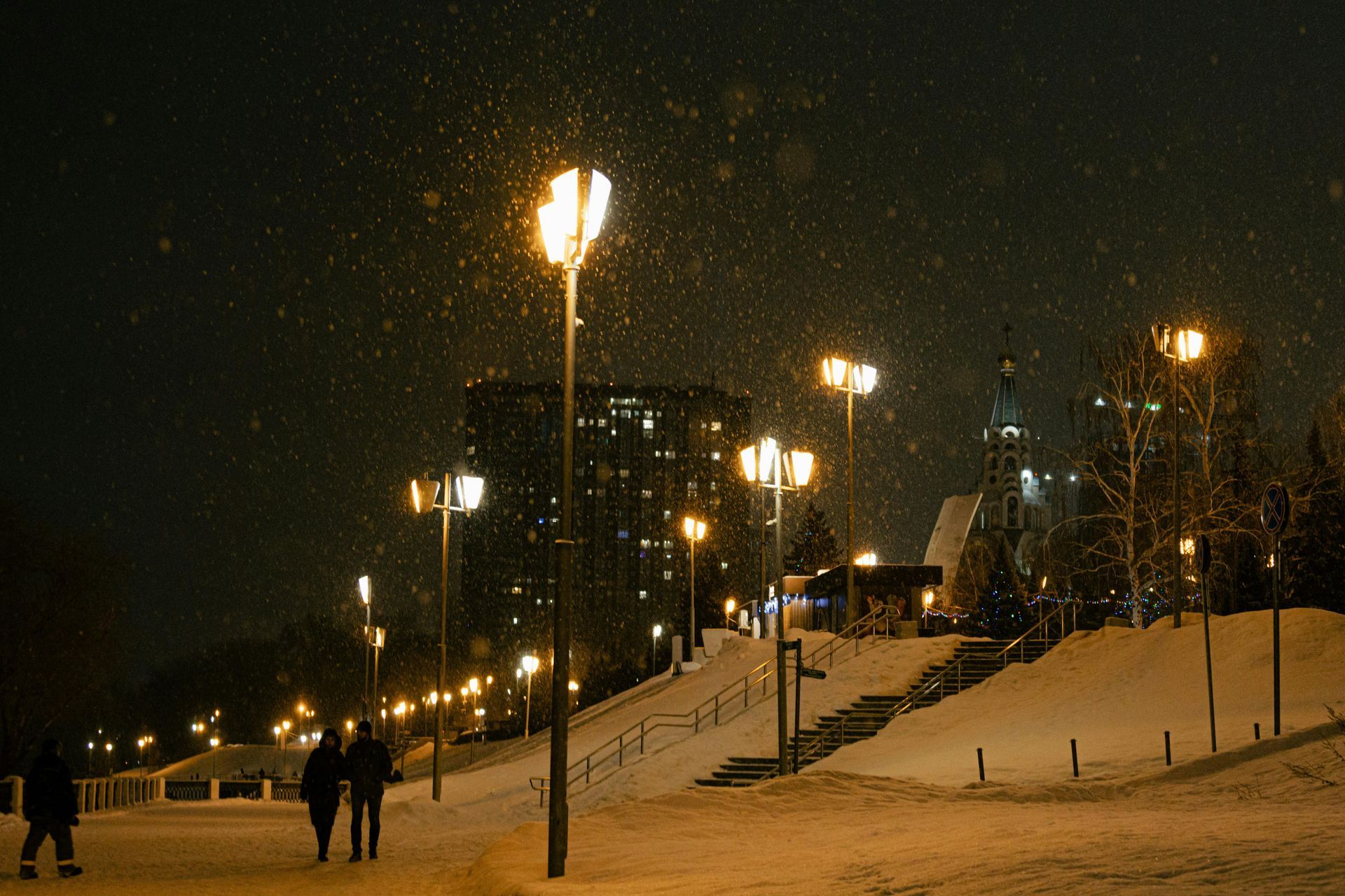 A couple is walking down a snowy sidewalk at night.