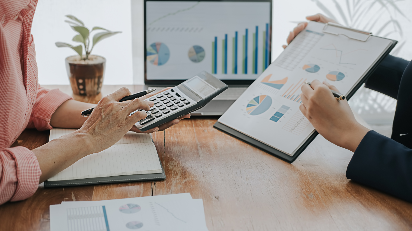 Two people reviewing financial data at a desk. One person uses a calculator, the other points at a chart.