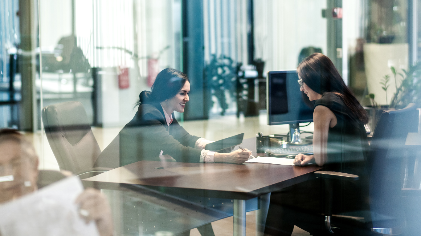 Two women in an office, seated at a desk, looking at documents. Through glass window.