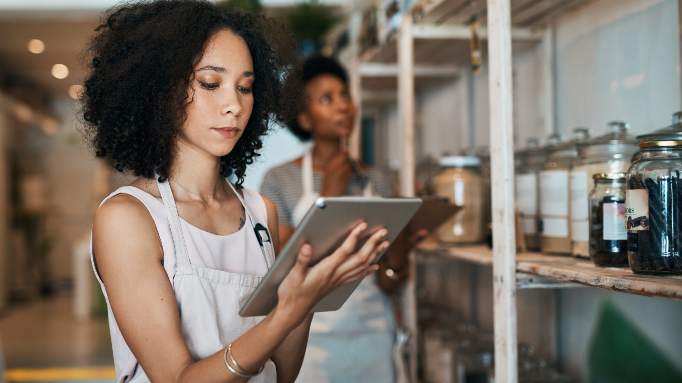 Woman in apron uses tablet, examining shelf with jars, another person in the background.