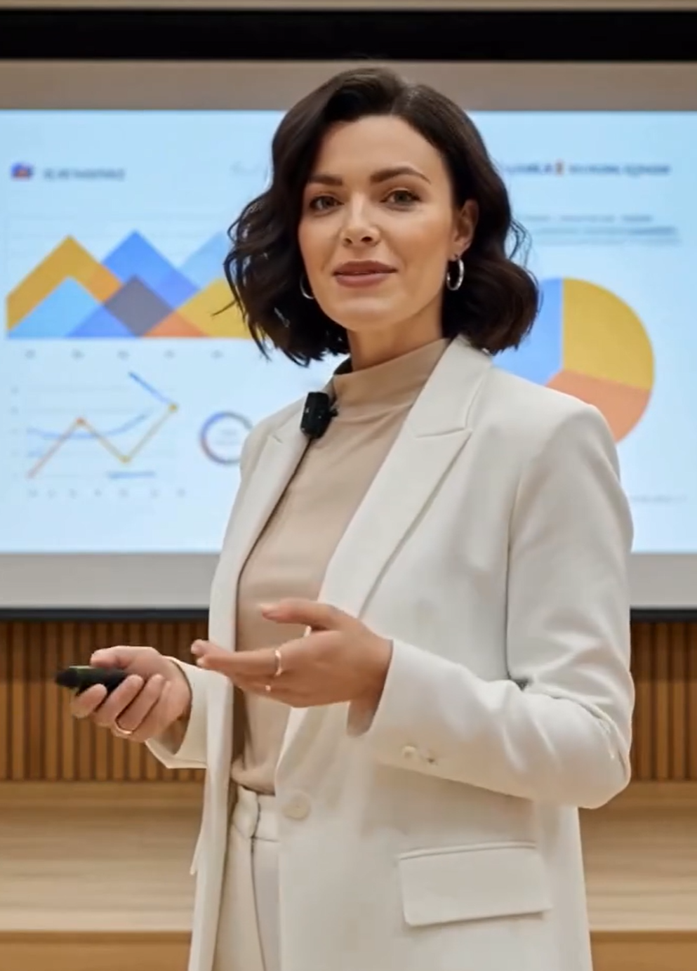 Woman in a white blazer presents data on a screen, holding a remote.