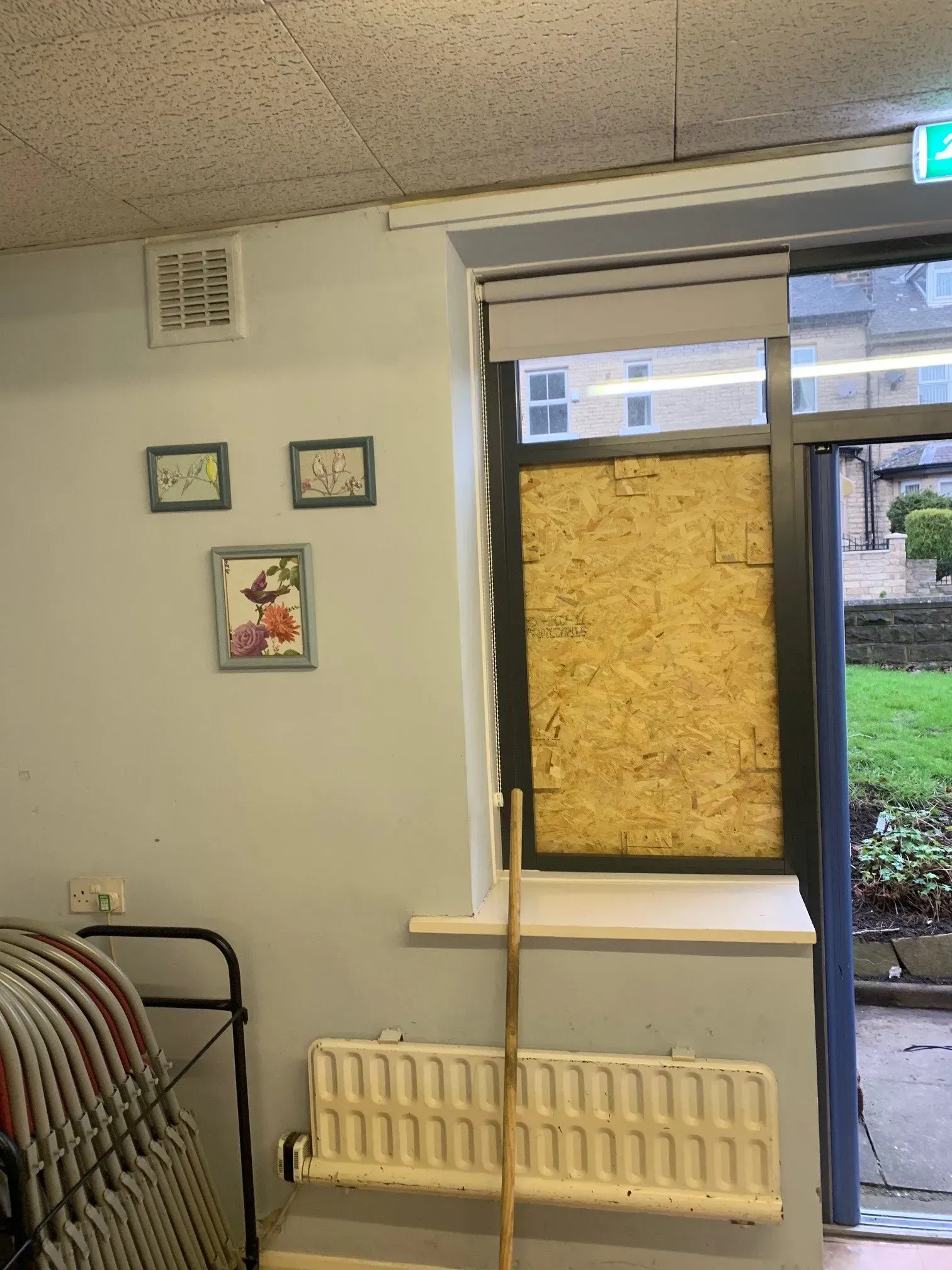 A boarded-up window next to a door. Three framed pictures hang above a radiator and stacked chairs.