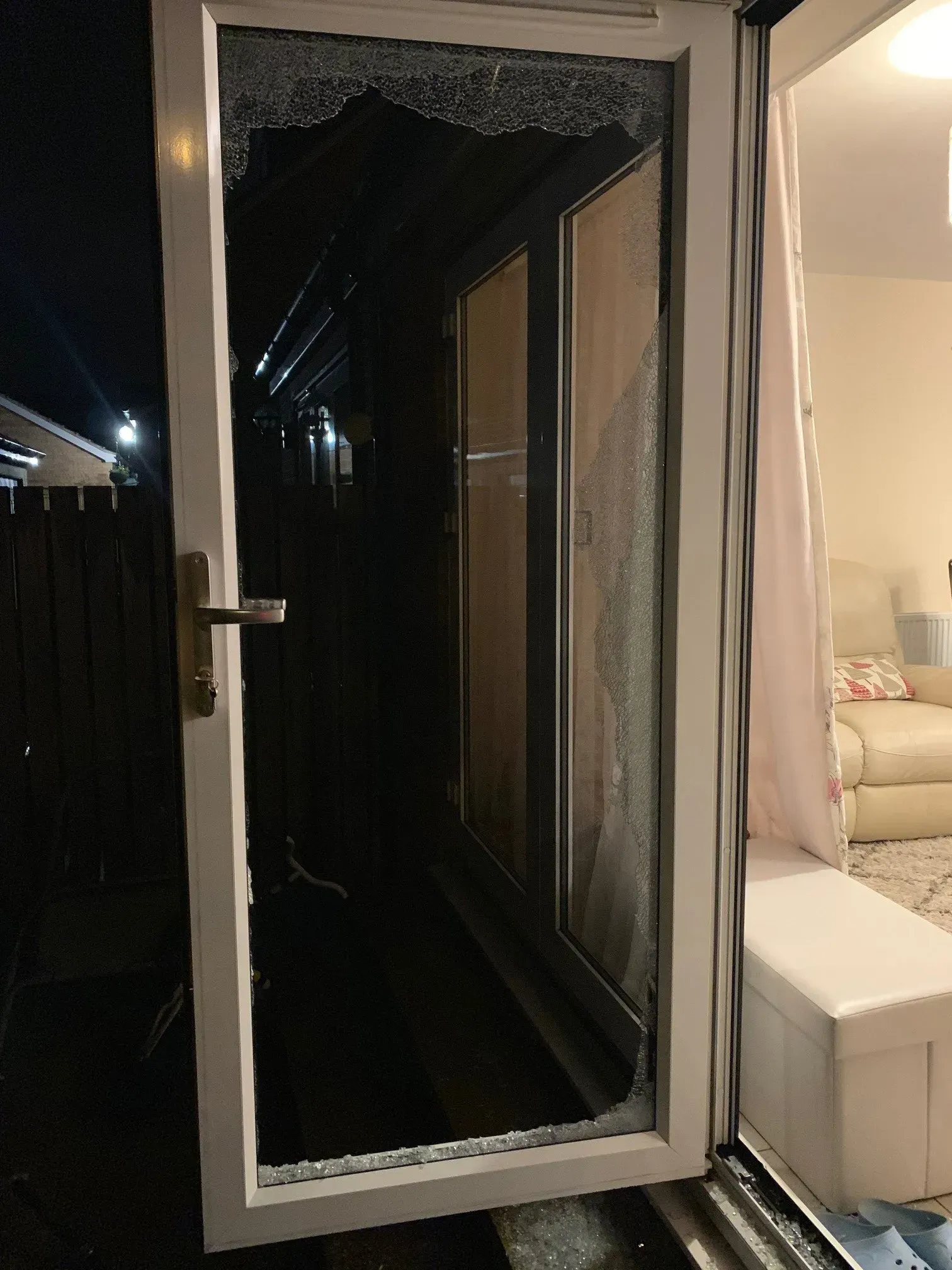 A white door with shattered glass. Inside a dimly lit home, a white bench with pink curtain is visible. Dark outside.