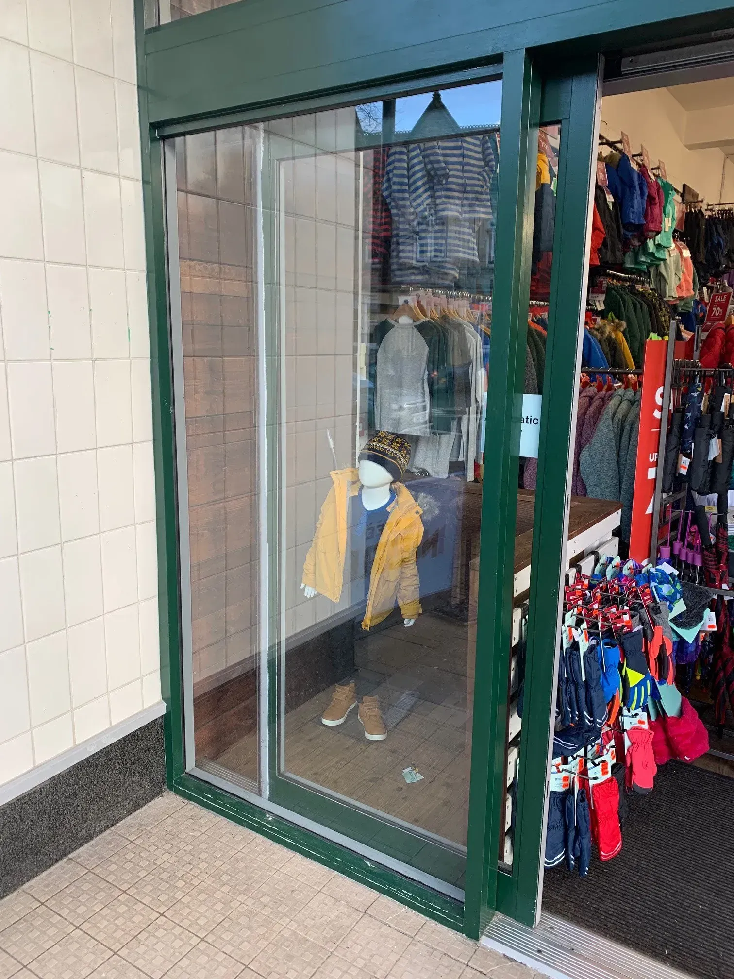 Glass store entrance with a mannequin wearing a yellow jacket and a partially visible rack of colorful clothing.