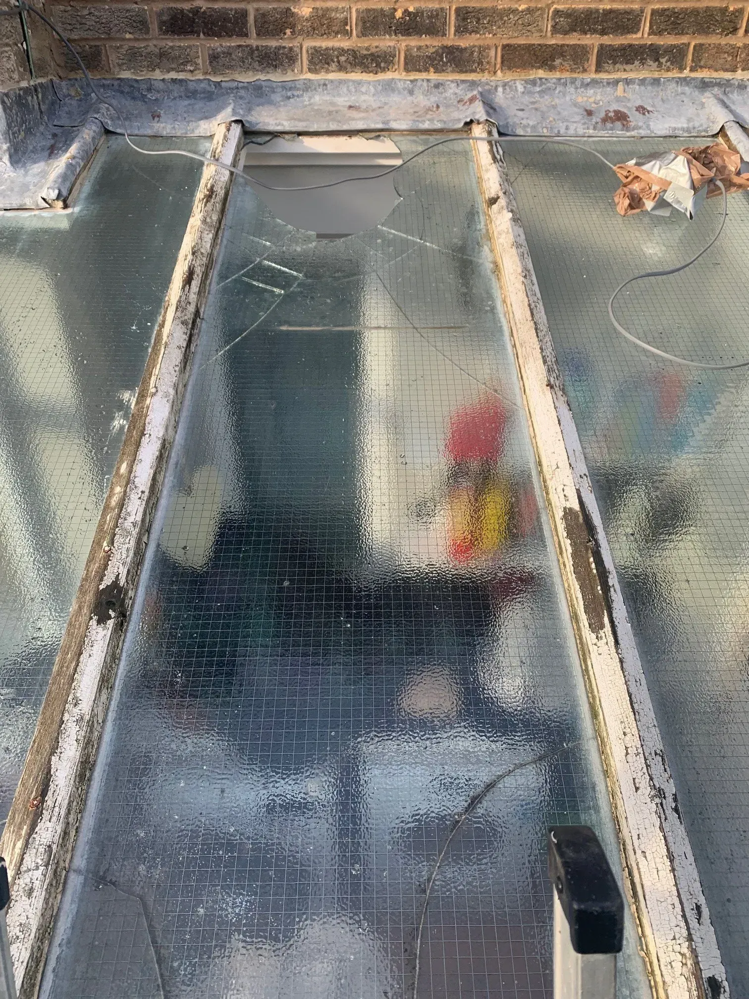 Cracked glass roof panel. Light reflects, revealing interior with brick wall and colorful objects.