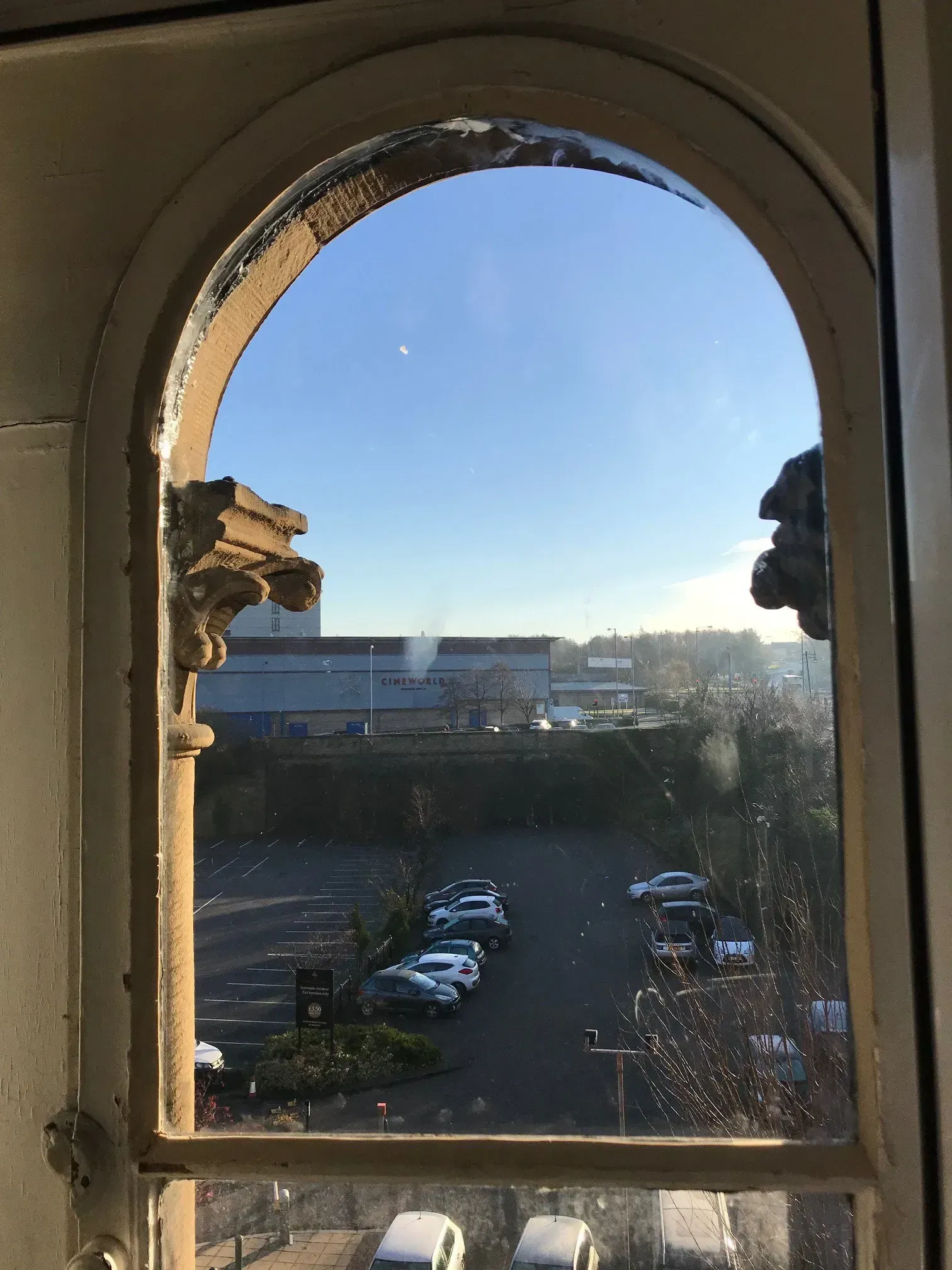 View through an arched window overlooking a parking lot and industrial buildings on a sunny day.