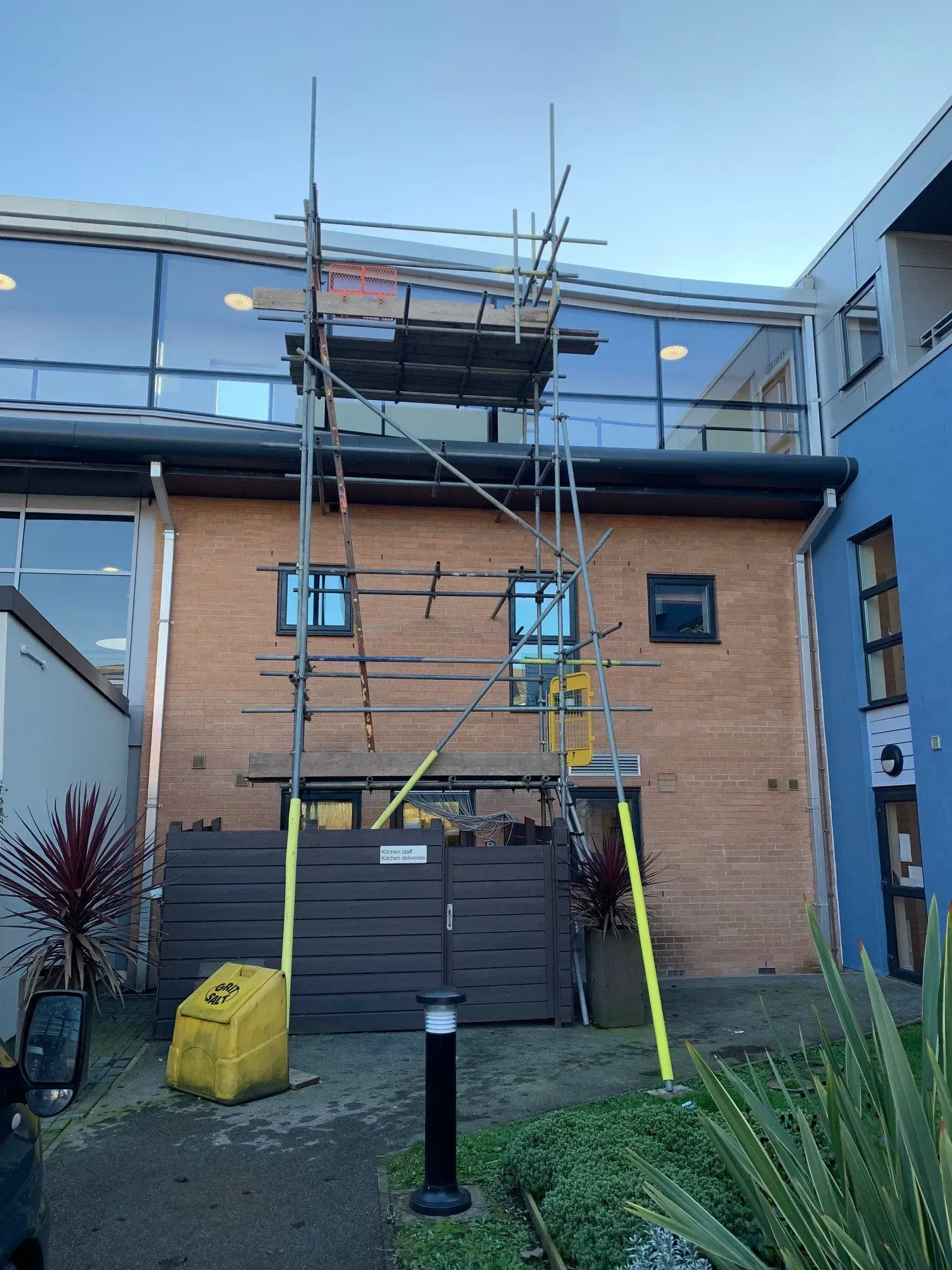 Scaffolding erected against a brick building with glass windows, a yellow generator, and blue/white walls.