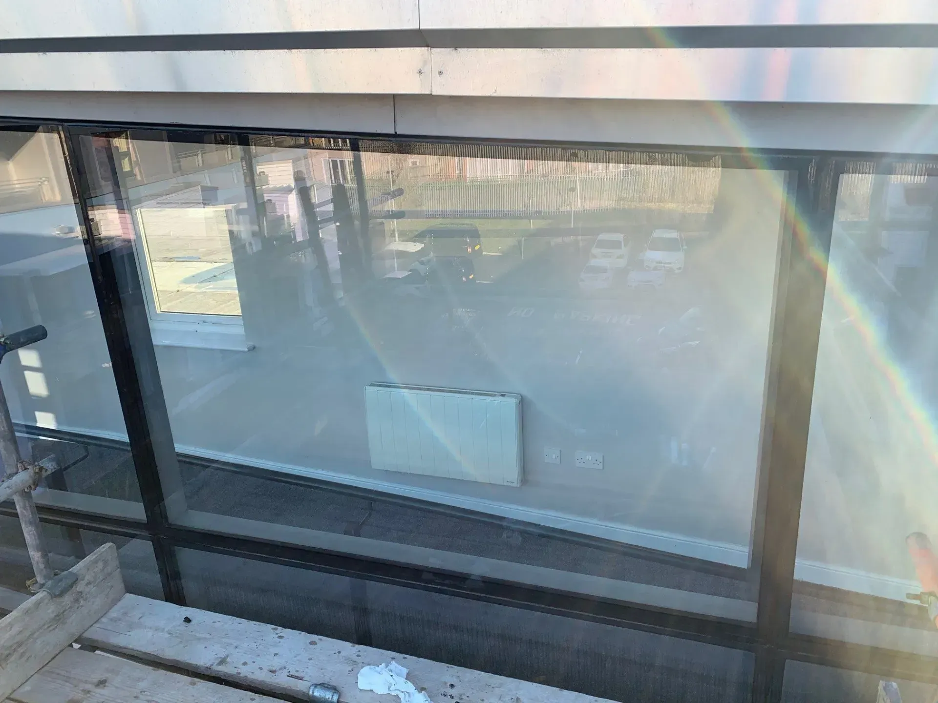 Exterior view through a large window, reflecting a sunny day. Black window frames, a white wall, and scaffolding visible.