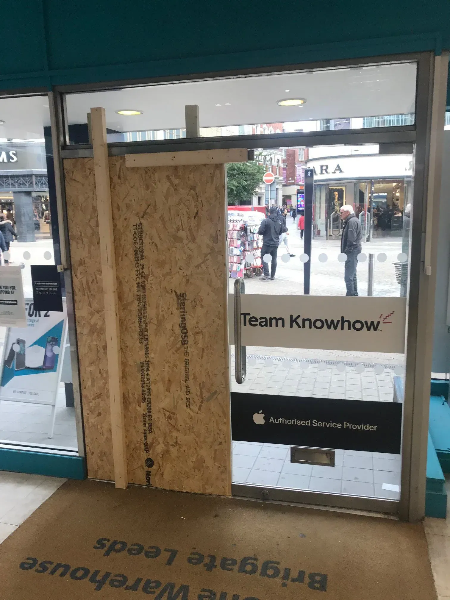 Boarded-up glass door at a store entrance, possibly damaged. A street scene is visible in the background.